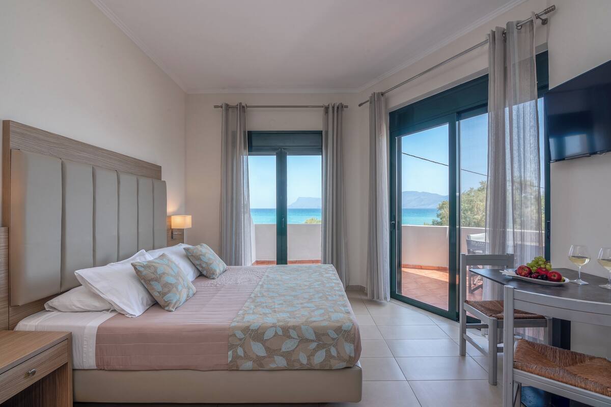 A hotel room with a bed, a table with fruit and wine glasses, and a balcony with a view of the sea.