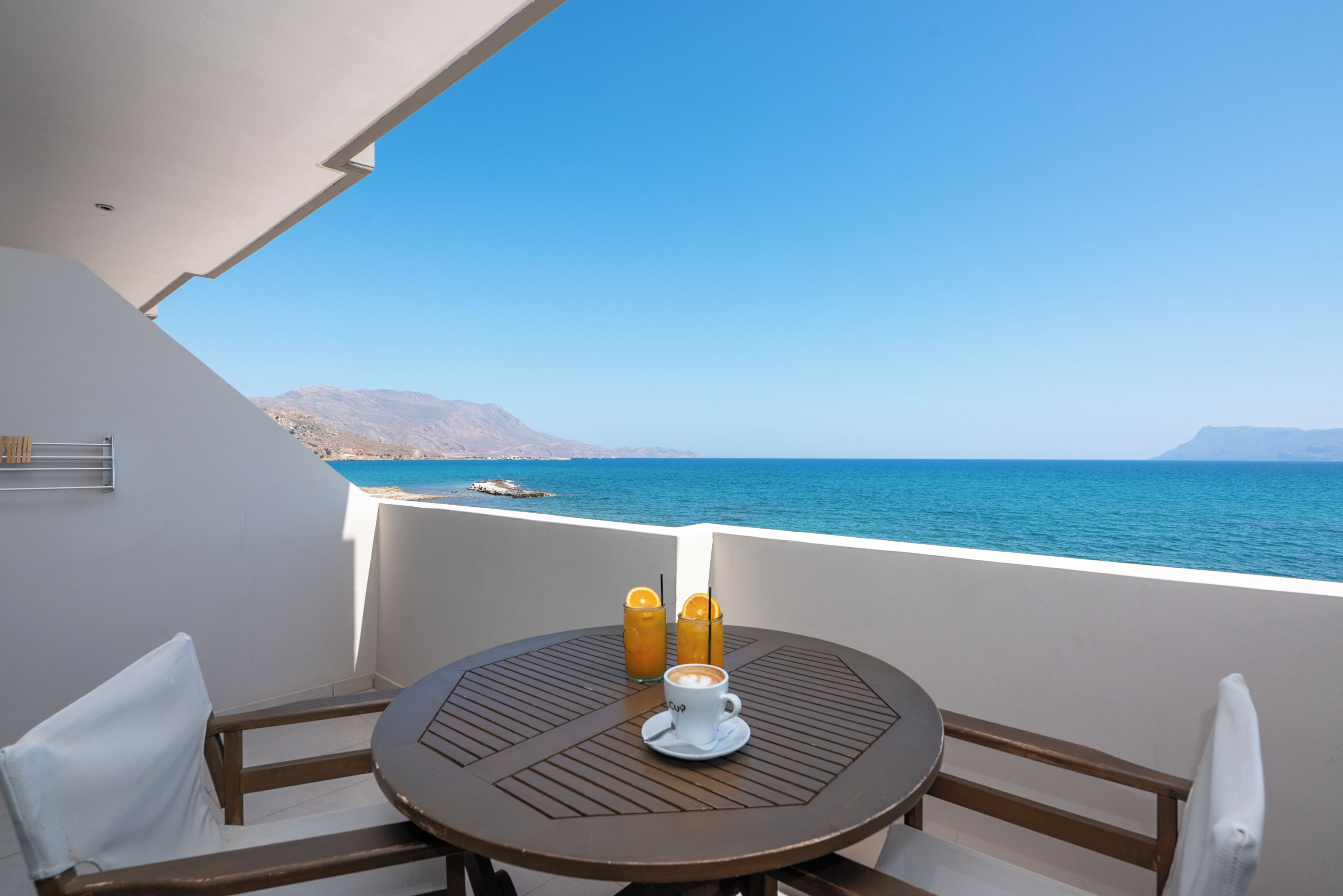A round table with a white cup and two drinks, overlooking a clear blue sea and distant mountains.
