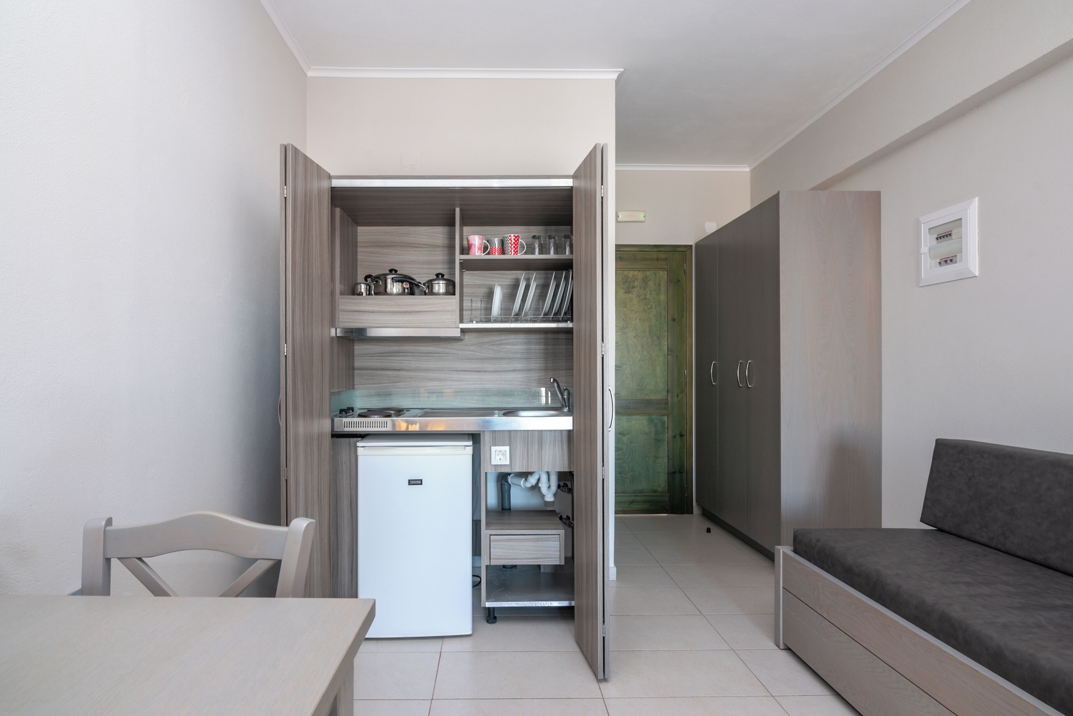 A compact kitchen with a refrigerator, sink, and open cabinets. There is a dining table with a chair and a sofa in the living area.