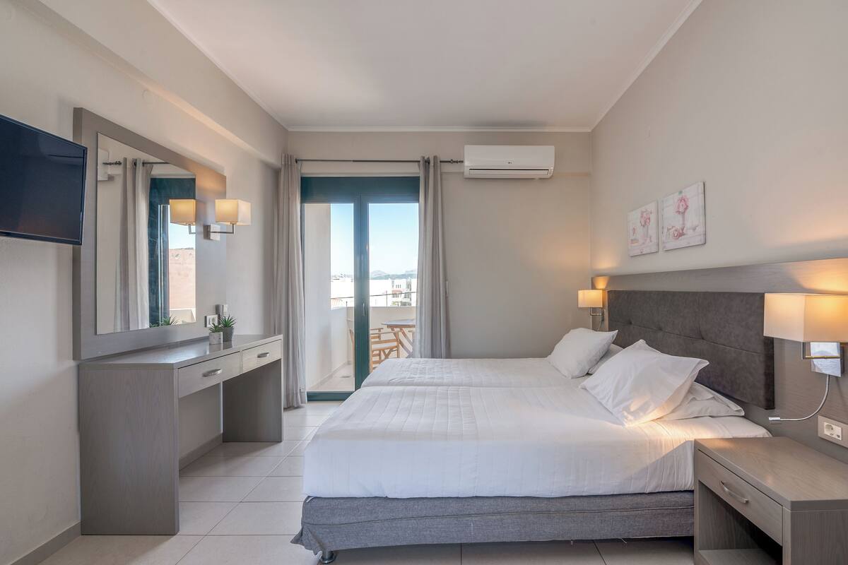 A modern hotel room with a large bed, a desk with a flat-screen TV, a balcony with a view, and an air conditioning unit.