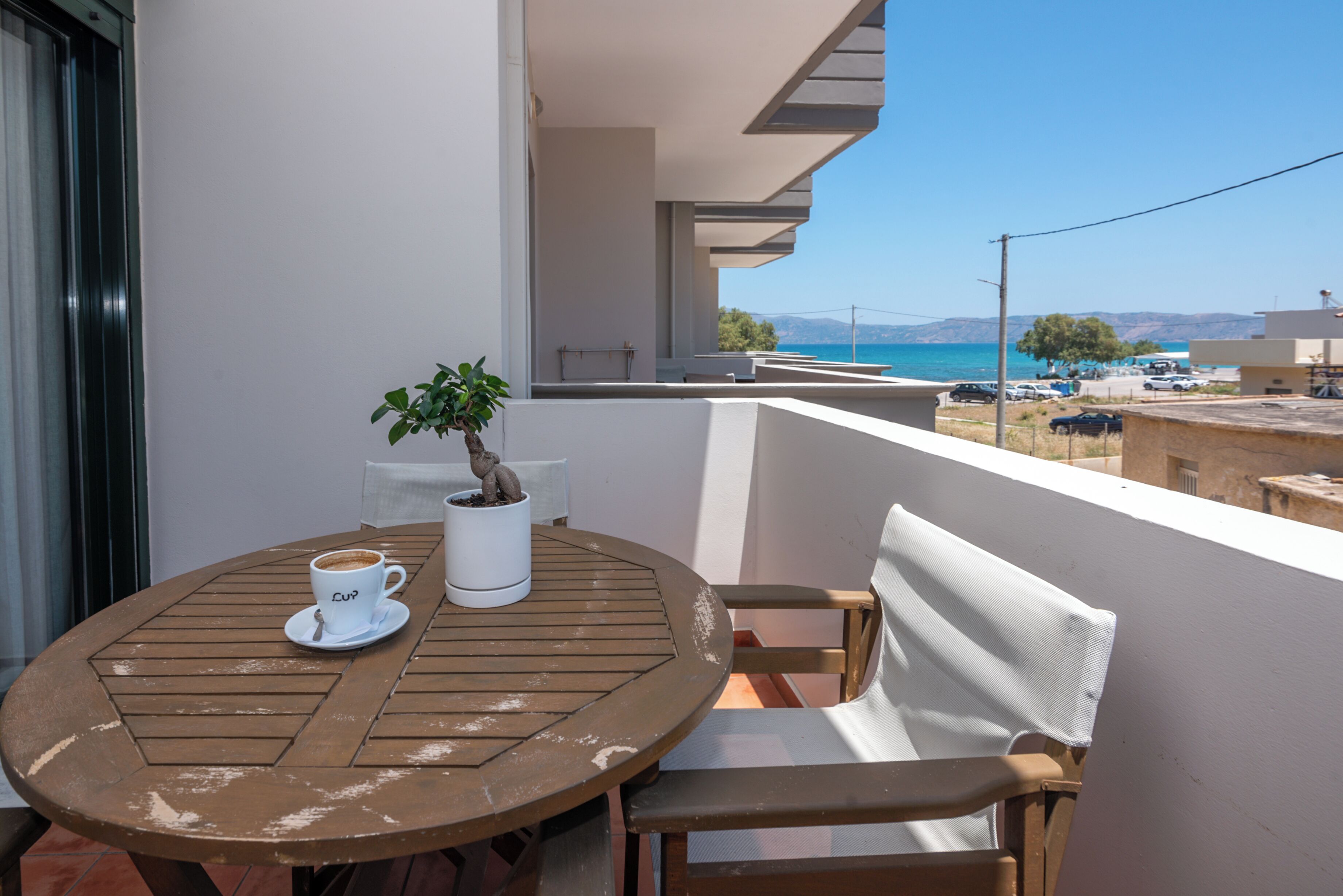 A round wooden table with a white cup and saucer, a potted plant, and a white vase on a balcony overlooking a coastal view.