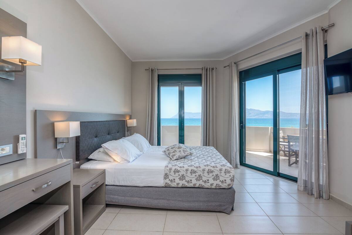 A hotel room with a bed, bedside table, lamp, and a view of the sea through a sliding door.