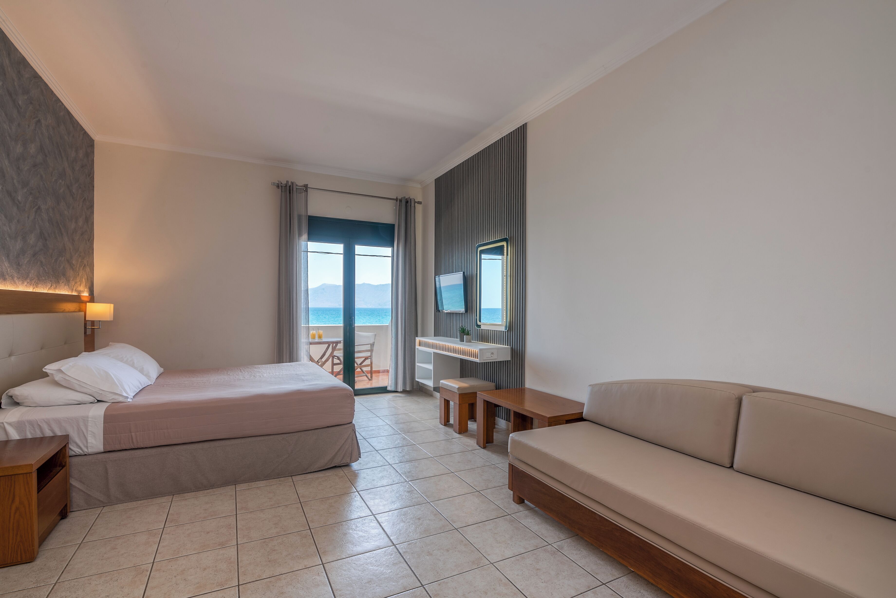 A hotel room with a bed, a sofa, a small table, a nightstand, a window with curtains, and a view of the sea.