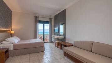 A hotel room with a bed, a sofa, a small table, a nightstand, a window with curtains, and a view of the sea.