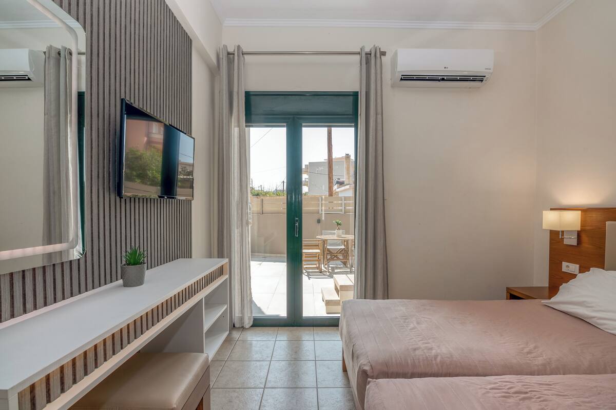 A hotel room with a bed, a TV mounted on the wall, a desk with a plant, and a sliding glass door leading to an outdoor area.
