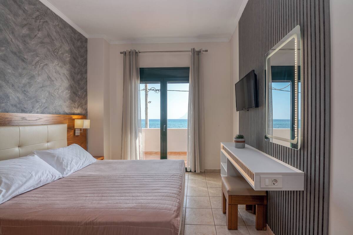 A modern hotel room with a bed, a TV, a vanity with a mirror, and a view of the sea.