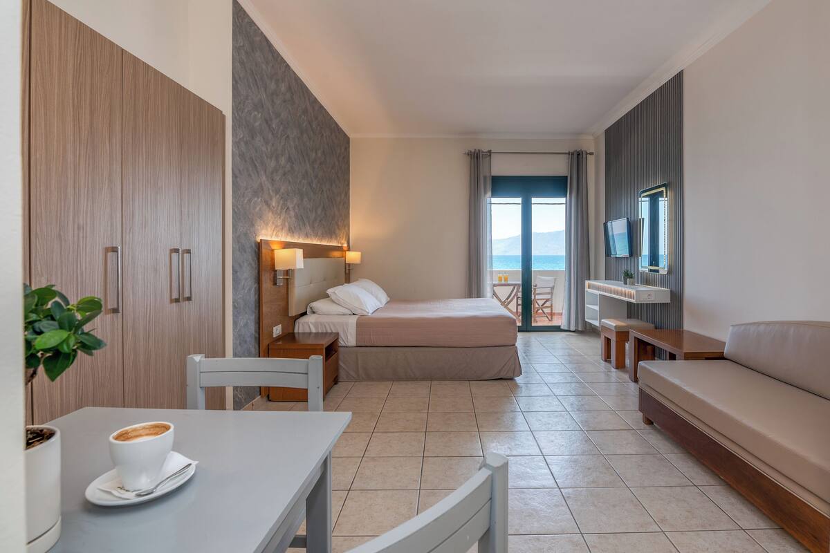 A modern hotel room with a bed, a desk with a cup of coffee, a TV, and a balcony with a view.