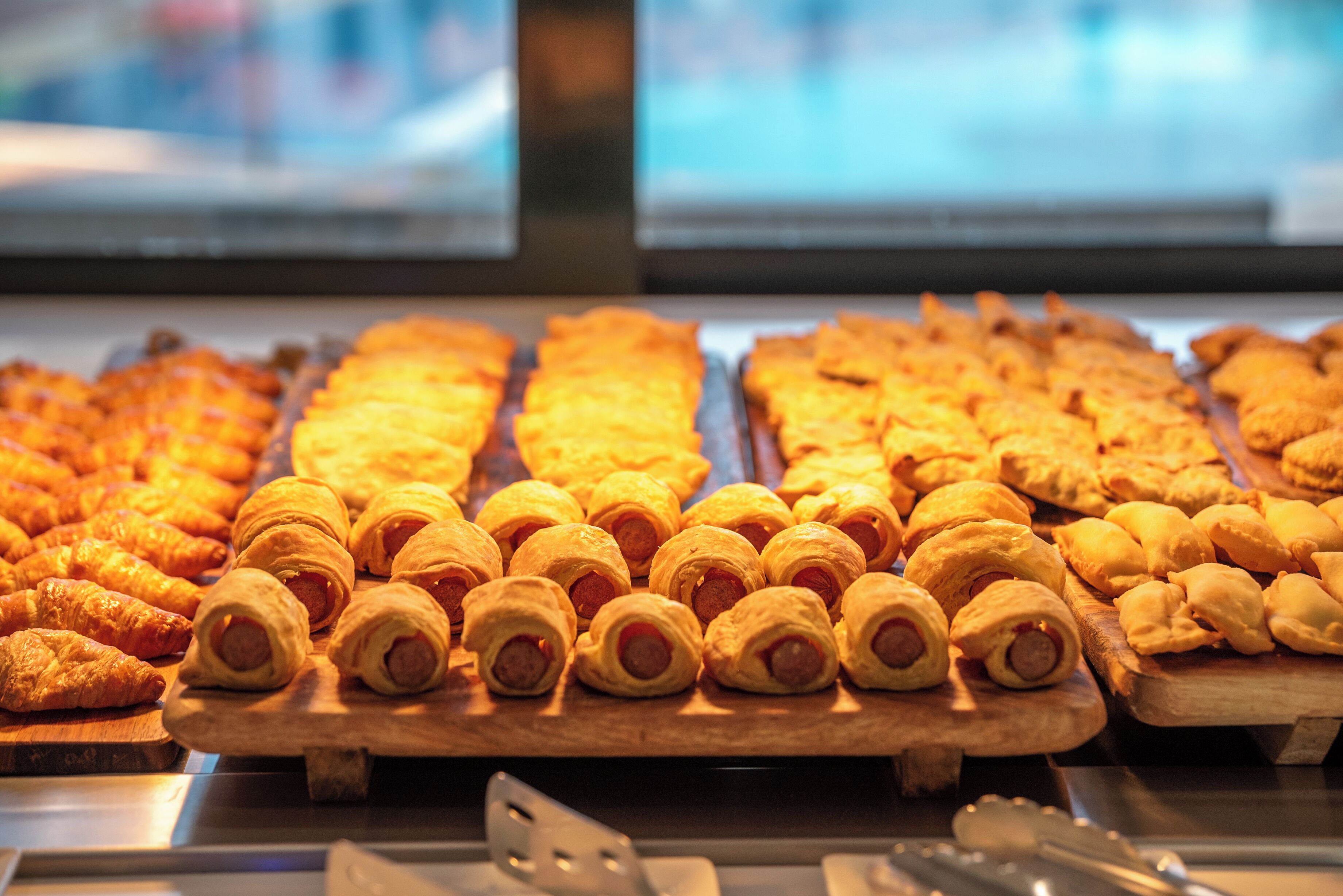 Una exhibición de diversos productos horneados, incluyendo croissants y rollos de salchicha, dispuestos sobre tablas de madera en un entorno de panadería.