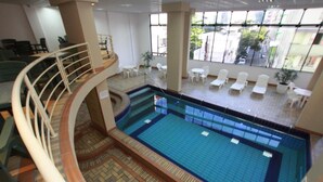 An indoor swimming pool with a tiled floor, a curved railing, and several lounge chairs around the pool area. - Hotel Blumenau (Balneário Camboriú)