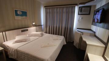 Quarto de hotel com cama, escrivaninha, televisão e janela com cortinas.