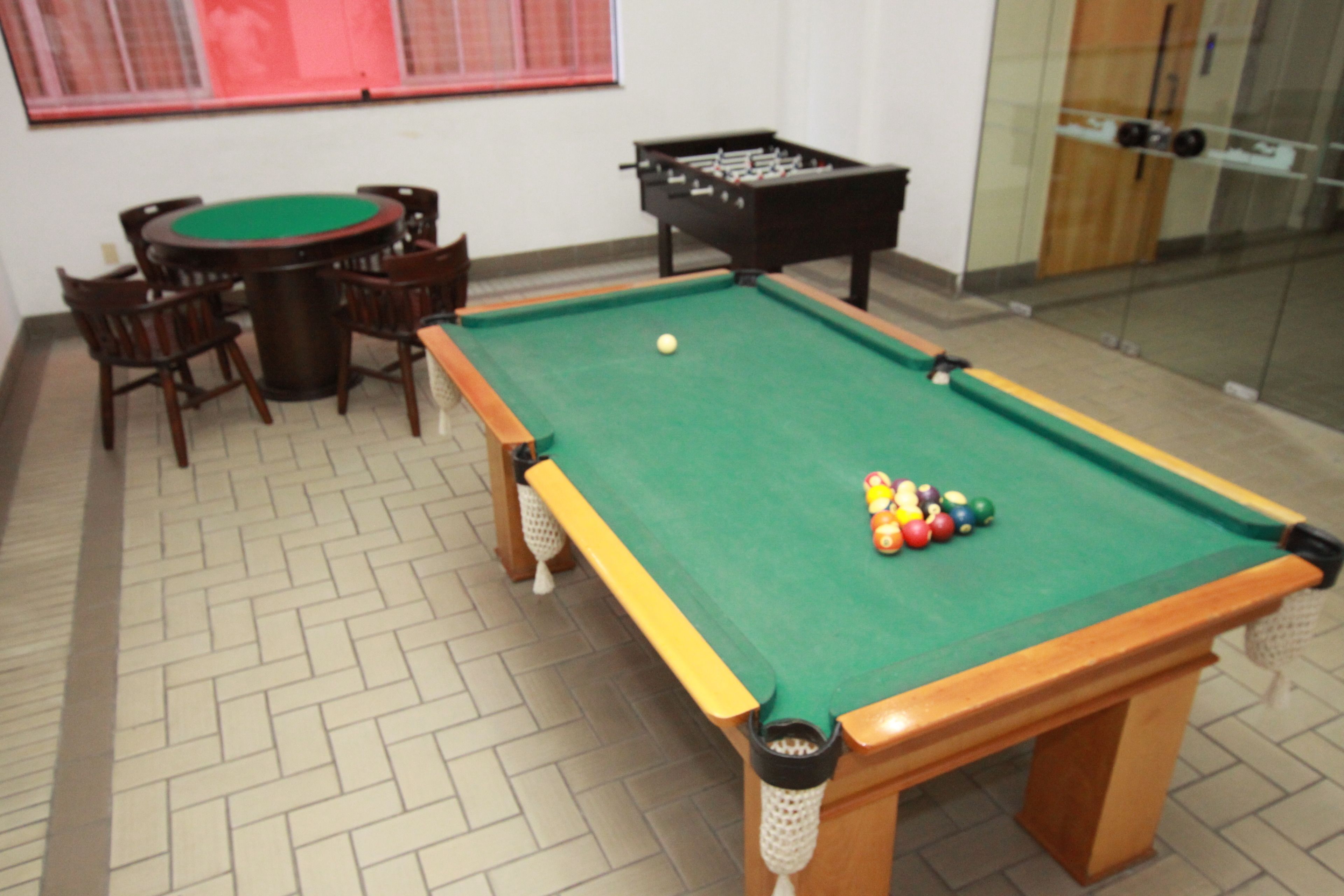 A pool table with a green felt surface, surrounded by wooden chairs, and a foosball table in the background.