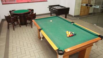 A pool table with a green felt surface, surrounded by wooden chairs, and a foosball table in the background.