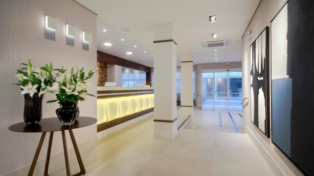 A hotel reception area with a modern design, featuring a reception desk, a waiting area, and a reception counter with a lighted panel. - Hotel Blumenau (Balneário Camboriú)