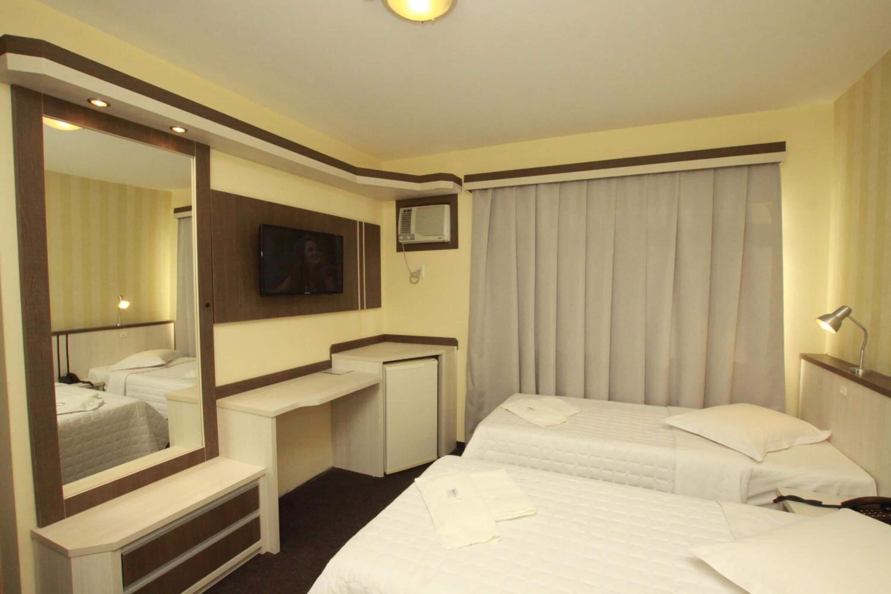 Habitación de hotel con dos camas, un escritorio, un espejo, un televisor y una ventana con cortinas.