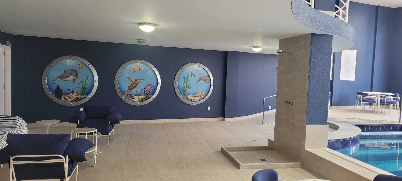 A pool area with three circular wall decorations depicting marine life, a seating area with chairs and tables, and a staircase leading to a balcony.