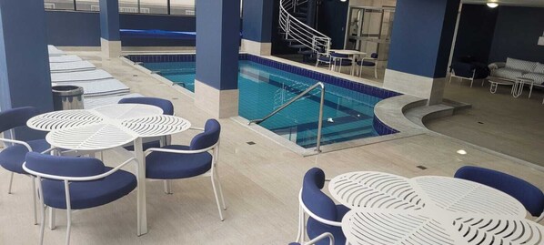 An indoor swimming pool area with multiple seating arrangements and a staircase leading to a higher level. - Hotel Blumenau (Balneário Camboriú)