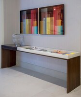 Un moderno mostrador de recepción con una vitrina de vidrio que contiene revistas, un jarrón de vidrio decorativo y dos pinturas abstractas de colores en la pared superior.