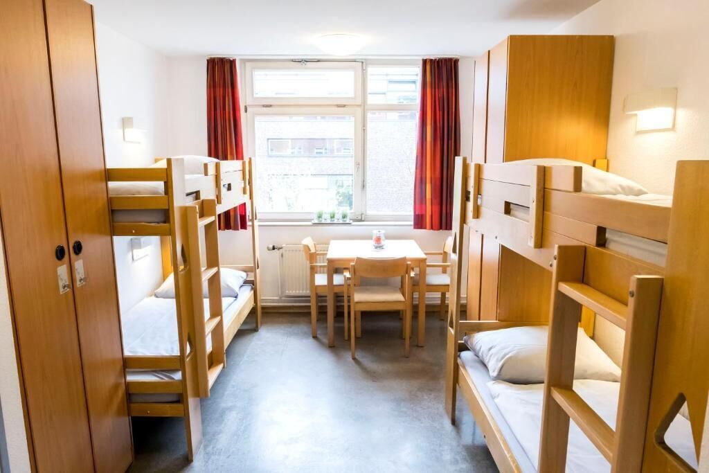 A room with bunk beds, a table, chairs, a window with curtains, and a radiator.
