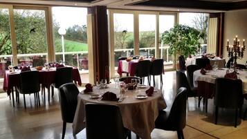 A dining area with round tables set for a meal, large windows offering a view of greenery, and a chandelier in the corner.