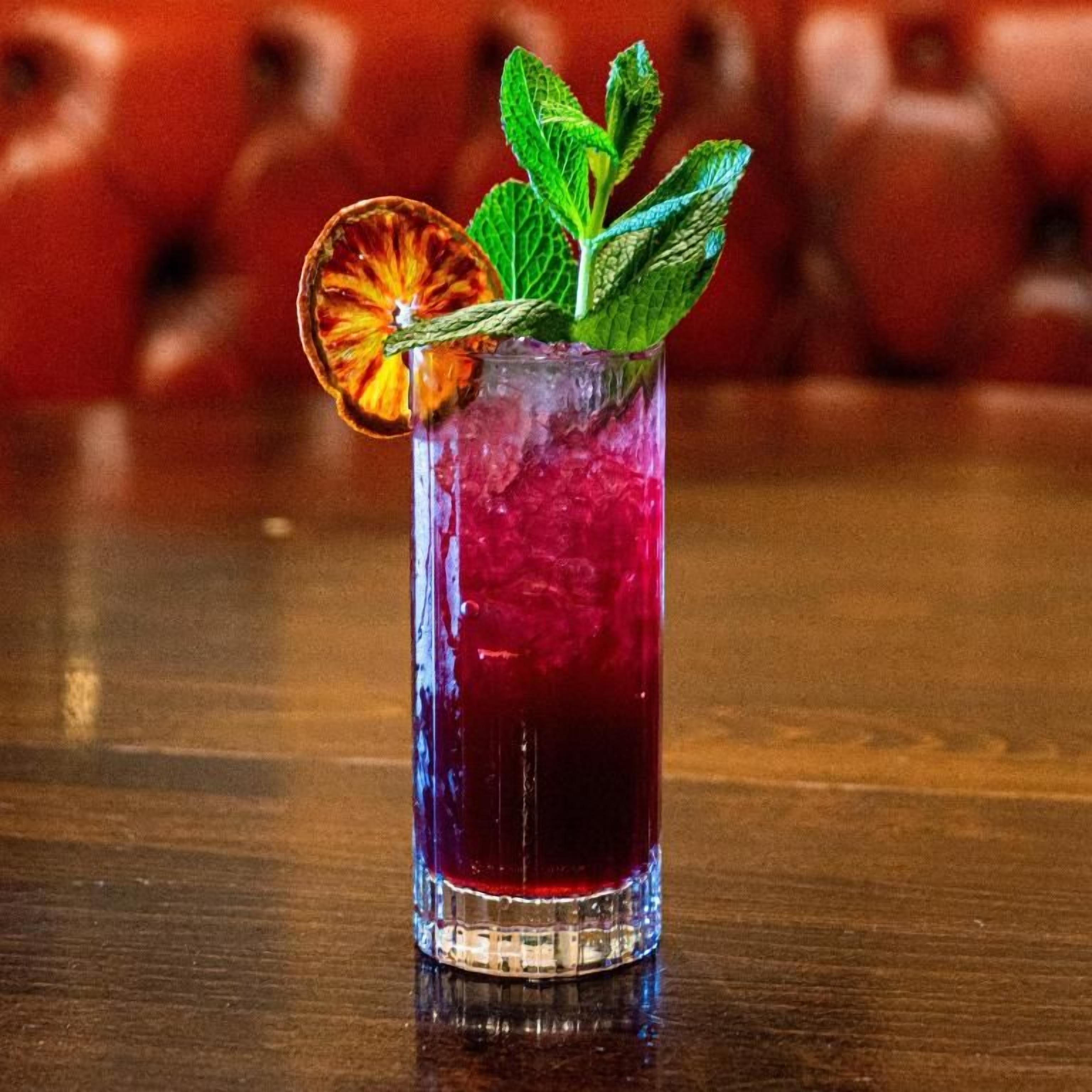 A tall glass with a red beverage, garnished with a dried orange slice and mint leaves, on a wooden surface.