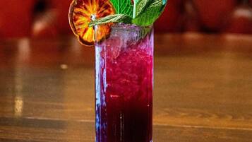 A tall glass with a red beverage, garnished with a dried orange slice and mint leaves, on a wooden surface.