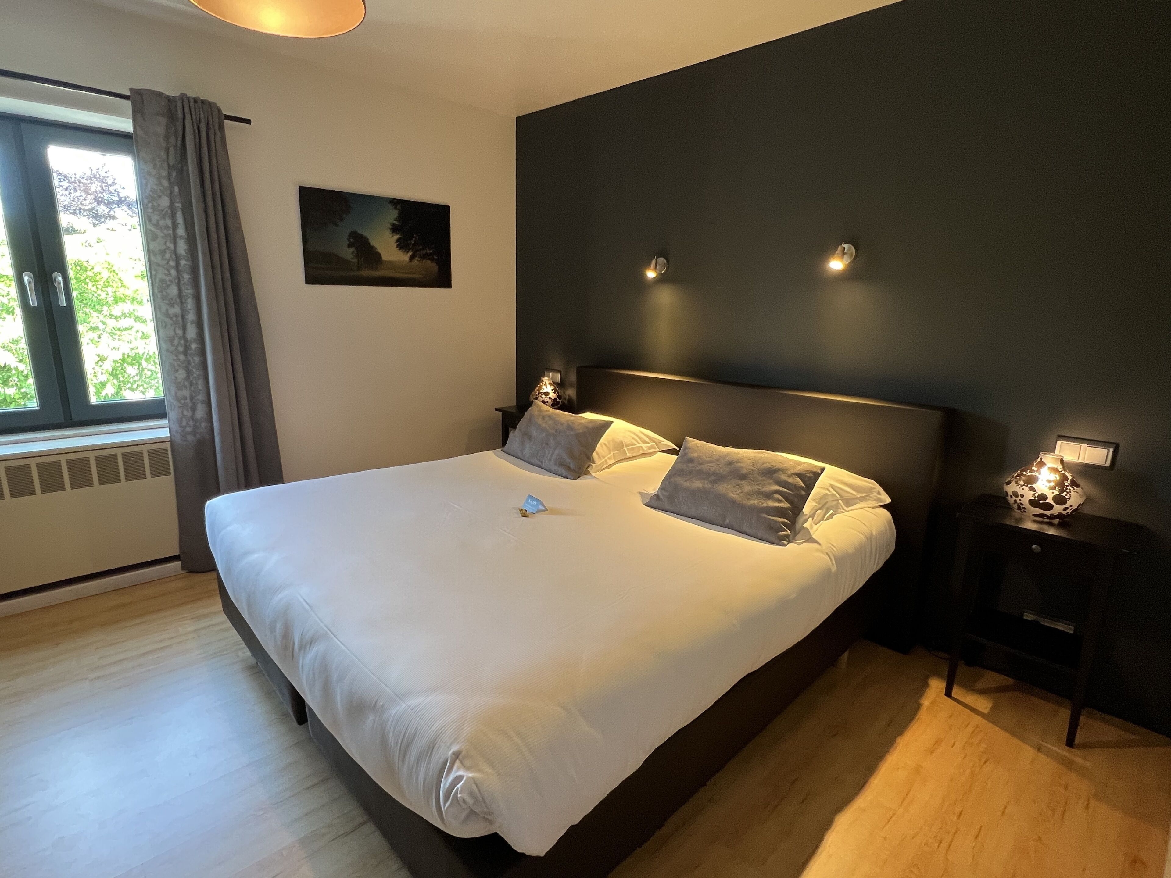 Uma cama grande com lençóis brancos e duas almofadas, uma mesa de cabeceira com abajur, um quadro na parede e uma janela com cortinas.