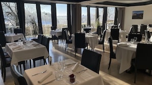 A restaurant with multiple tables set for dining, large windows offering a view of the outdoors, and a painting on the wall.