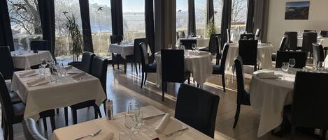 A restaurant with multiple tables set for dining, large windows offering a view of the outdoors, and a painting on the wall.