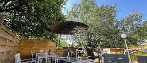 Un área de comedor al aire libre con mesas y sillas, una valla de madera y un árbol grande que da sombra.