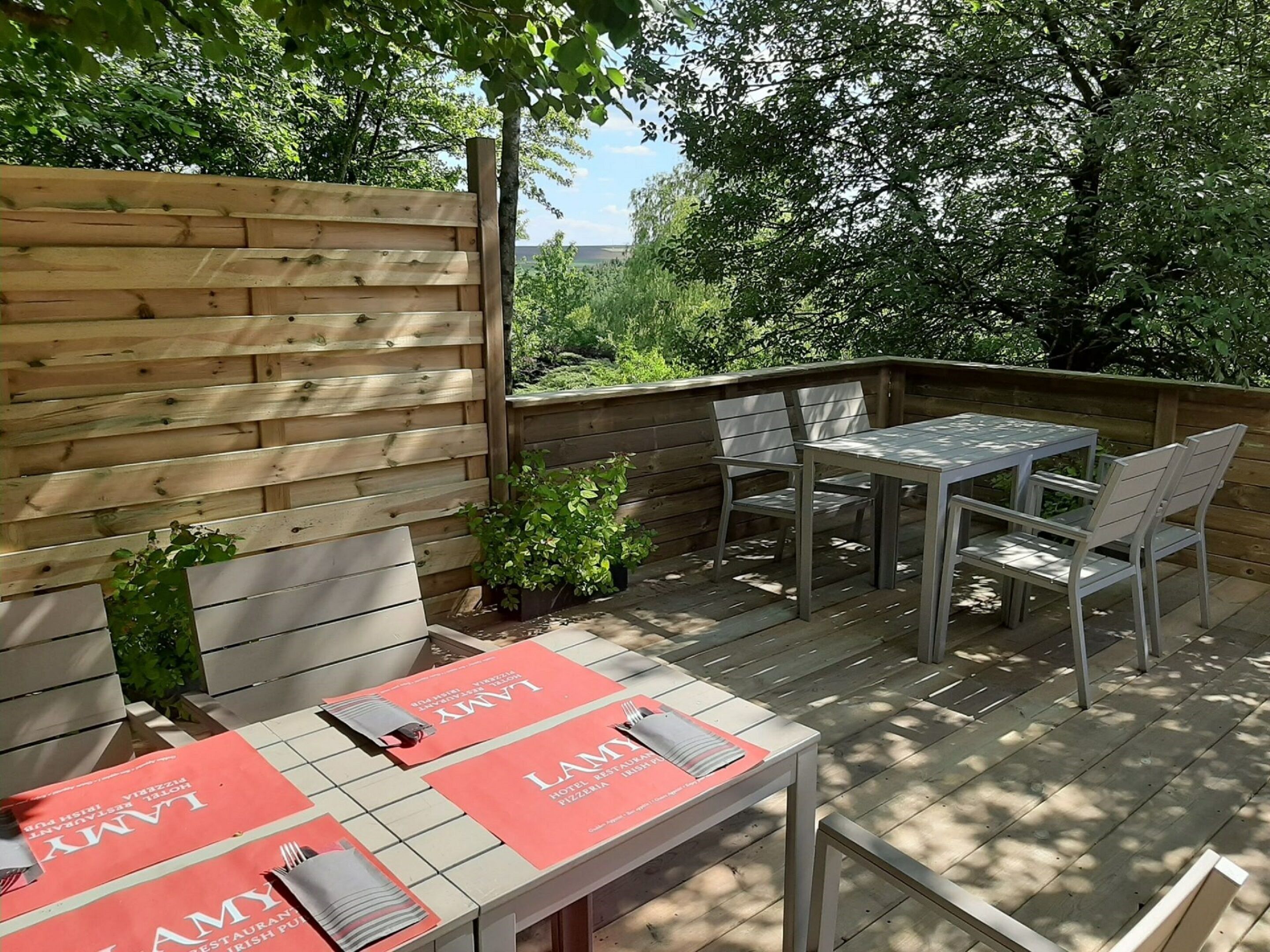 Eine Terrasse im Freien mit Holzdeck, einem Metalltisch und -stühlen sowie einer roten Tischmatte mit dem Namen "LAMY", das auf ein Restaurant hindeutet.
