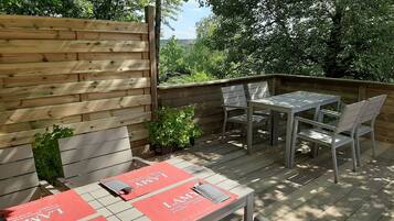 Een terras buiten met een houten terras, een metalen tafel en stoelen, en een rode placemat met de naam 'LAMY', wat duidt op een restaurant.
