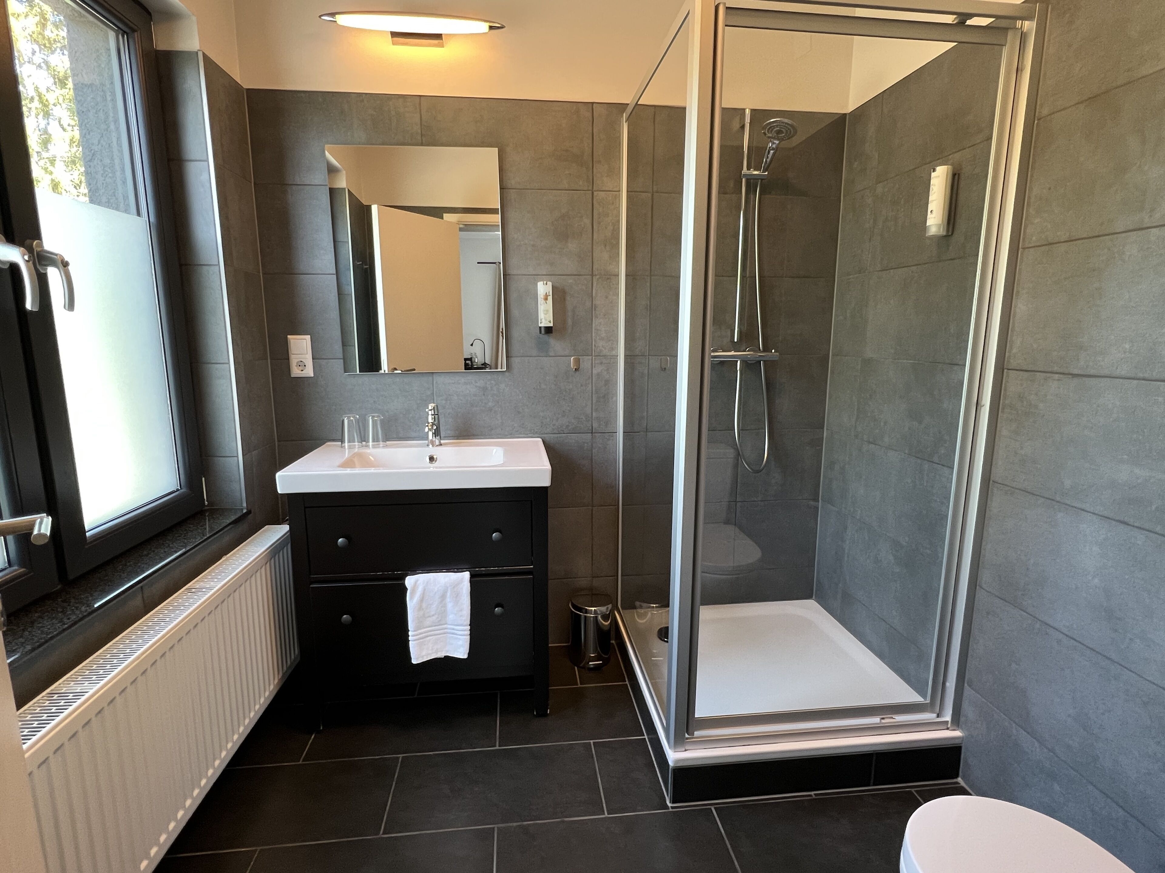Un baño moderno con ducha de vidrio, lavamanos blanco y tocador de tonos oscuros.