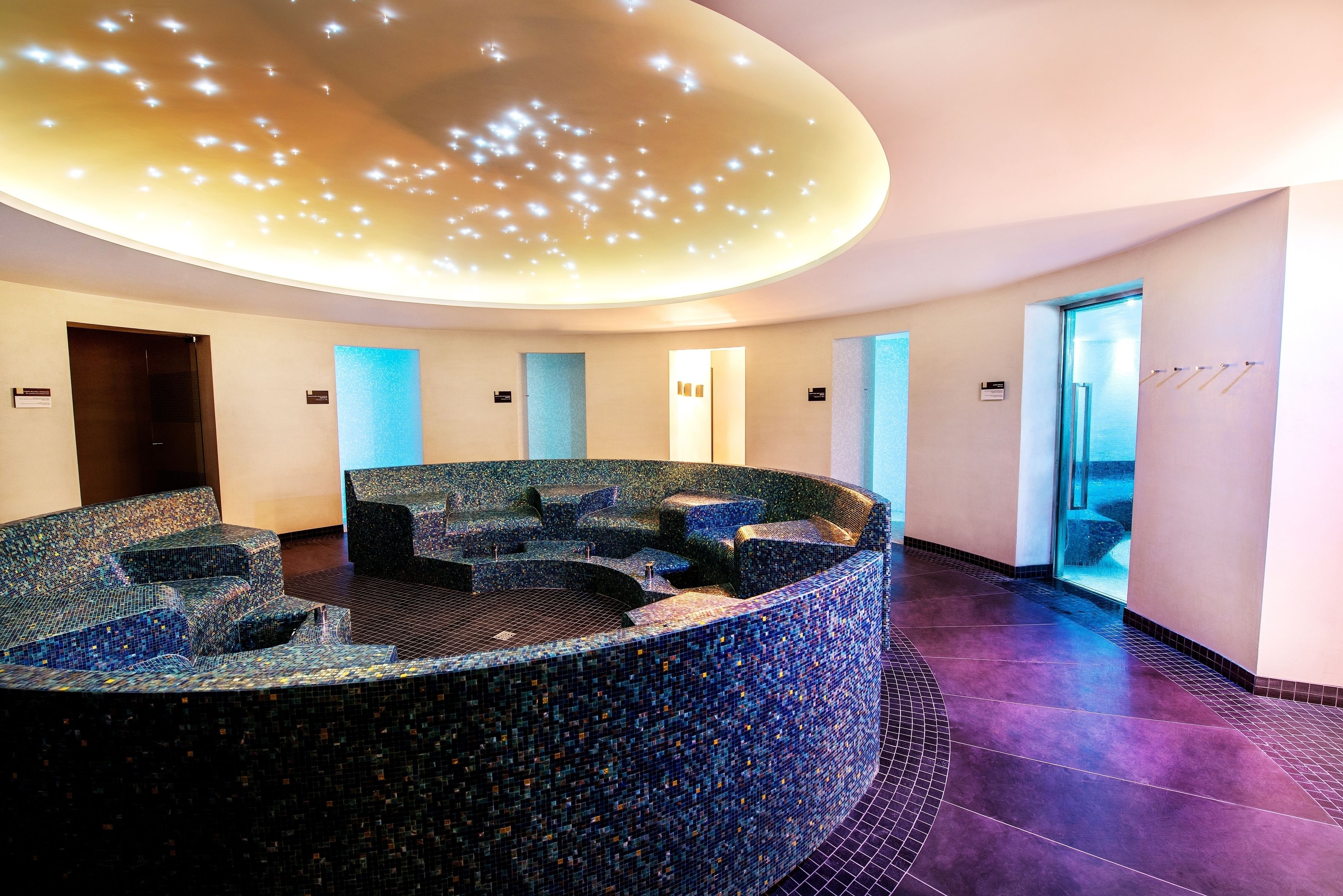 Un espace circulaire aménagé avec des sièges, un sol en mosaïque, un plafond doté de luminaires encastrés et une porte donnant sur l’extérieur.
