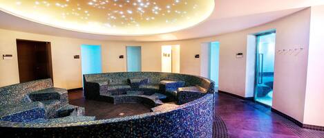 Un espace circulaire aménagé avec des sièges, un sol en mosaïque, un plafond doté de luminaires encastrés et une porte donnant sur l’extérieur.