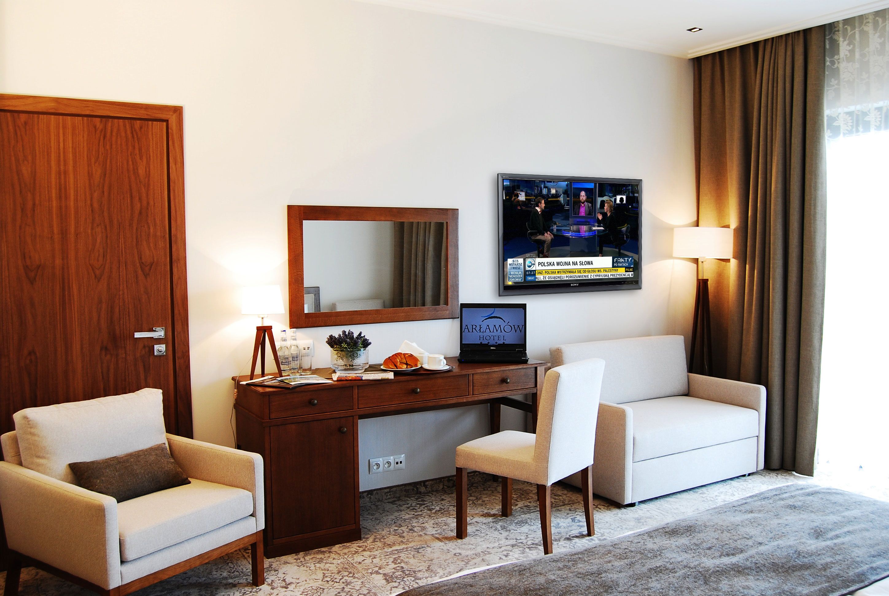 Quarto de hotel com secretária de madeira, televisão, sofá e uma cadeira.