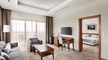 Habitación de hotel con sofá, escritorio, televisión, cama y un ventanal grande con cortinas.