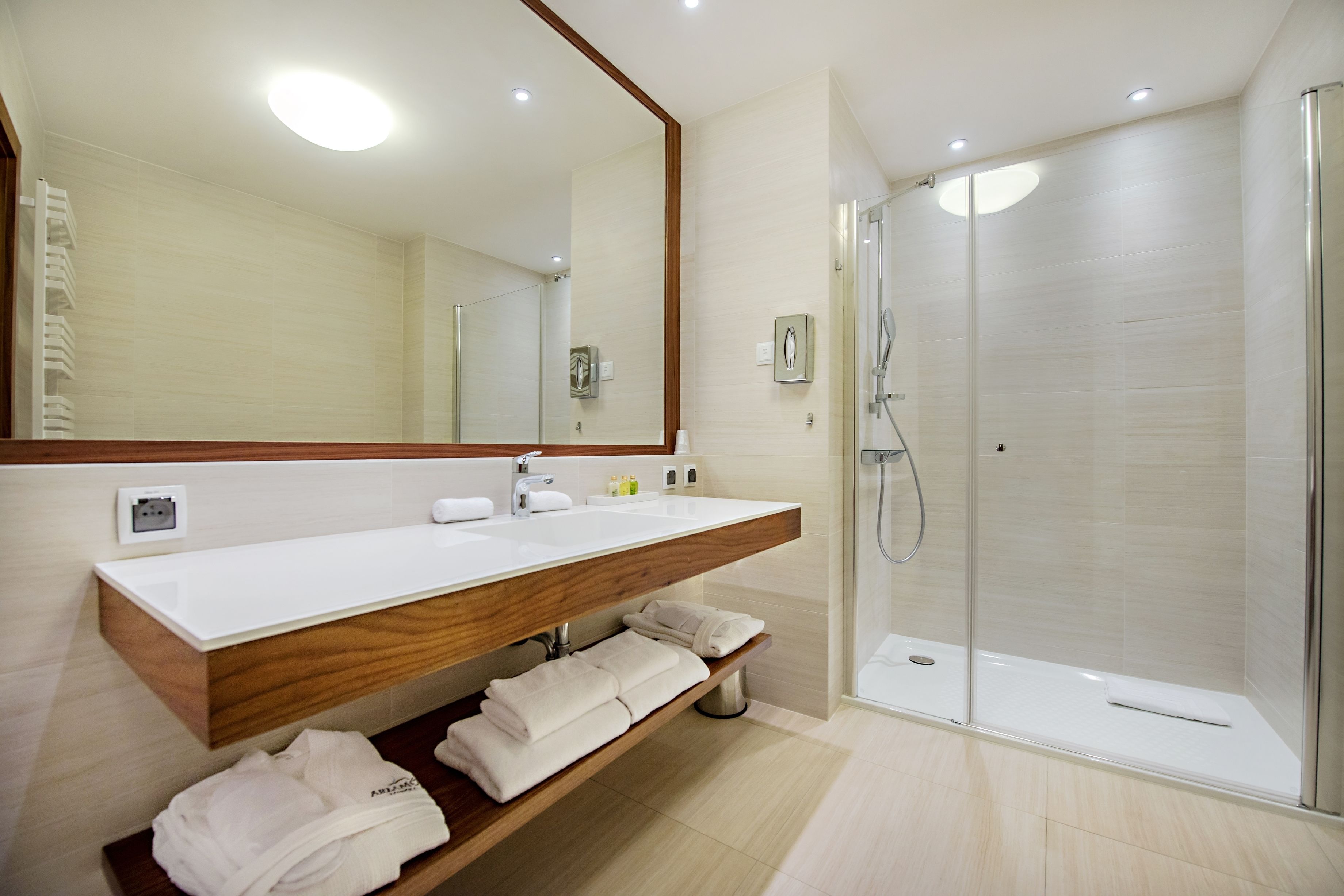 Un baño moderno con tocador de madera, un espejo grande, cabina de ducha con mampara de vidrio y toallas dobladas ordenadamente en un estante.