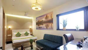A hotel room with a bed, a sofa, a dining table, a ceiling fan, a chandelier, a window with a plant, and a painting of a famous monument. - Hotel Taj Inn (Agra)