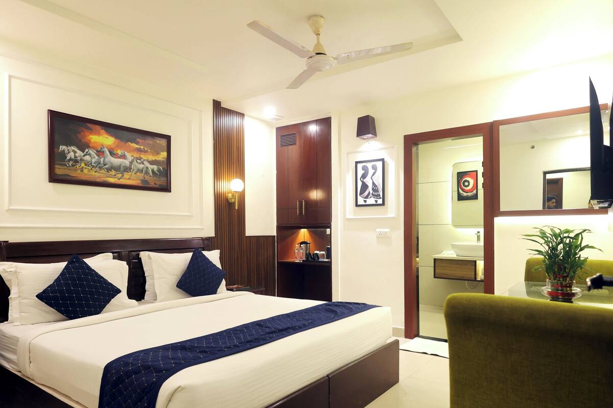 A hotel room with a bed, a ceiling fan, a painting, a mirror, and a potted plant.