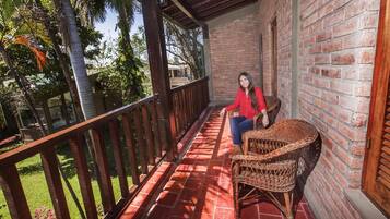 A covered balcony with a brick wall, a wicker chair, and a view of greenery.
