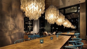 A modern dining area with long wooden tables, blue chairs, and large, ornate chandeliers. - Motel One Rostock (Rostock)