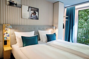 A hotel room with a large bed, two pillows, a headboard, a bedside table with a lamp, and a window with curtains.