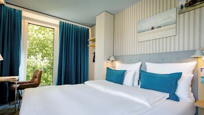 A hotel room with a large bed, a desk, and a chair. There is a window with curtains, a wall-mounted lamp, and a painting on the wall. - Motel One Rostock (Rostock)