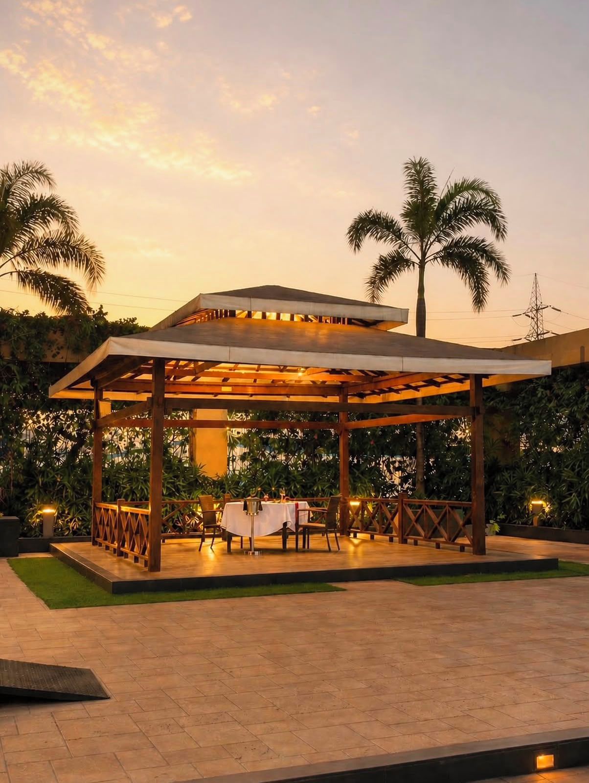 A covered outdoor seating area with tables and chairs, surrounded by greenery and palm trees.