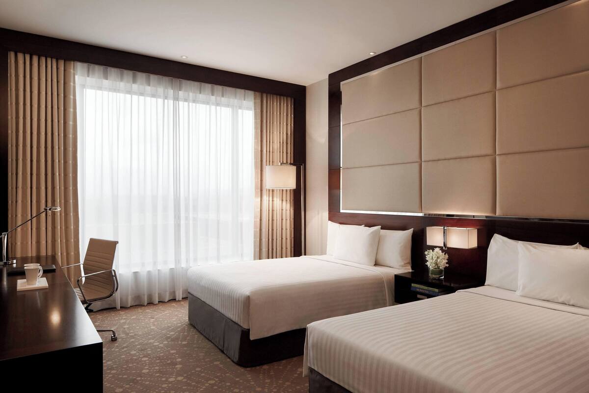A hotel room with two beds, a desk, a chair, and a large window with curtains.