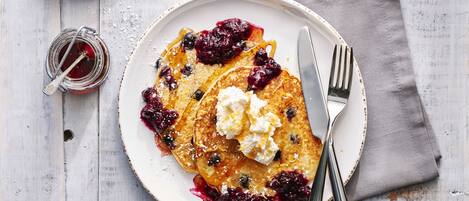 A plate of pancakes with berries and whipped cream, a jar of syrup, and a cup of coffee on a wooden table.