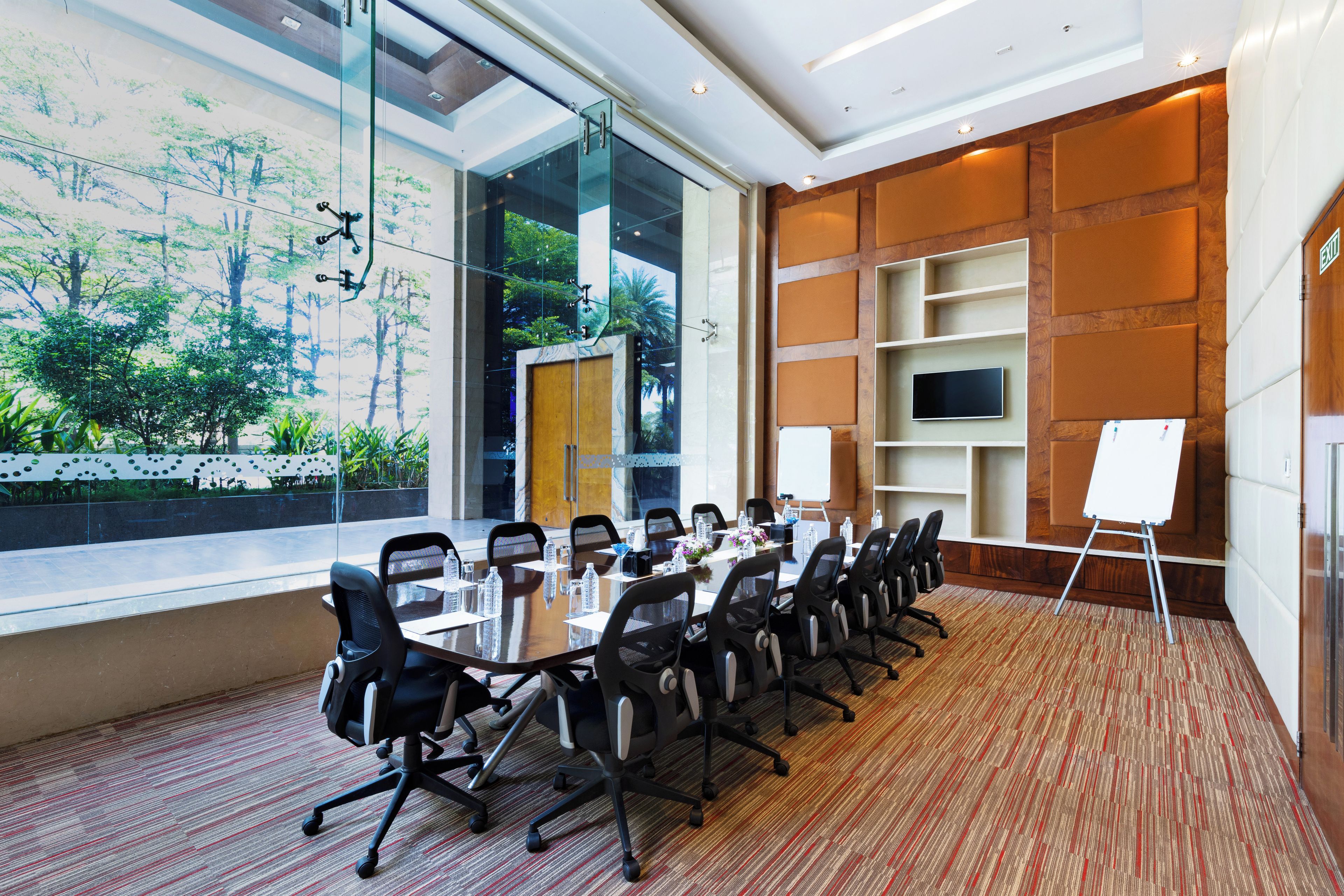 A conference room with a large table, chairs, a whiteboard, and a view of greenery through the windows.