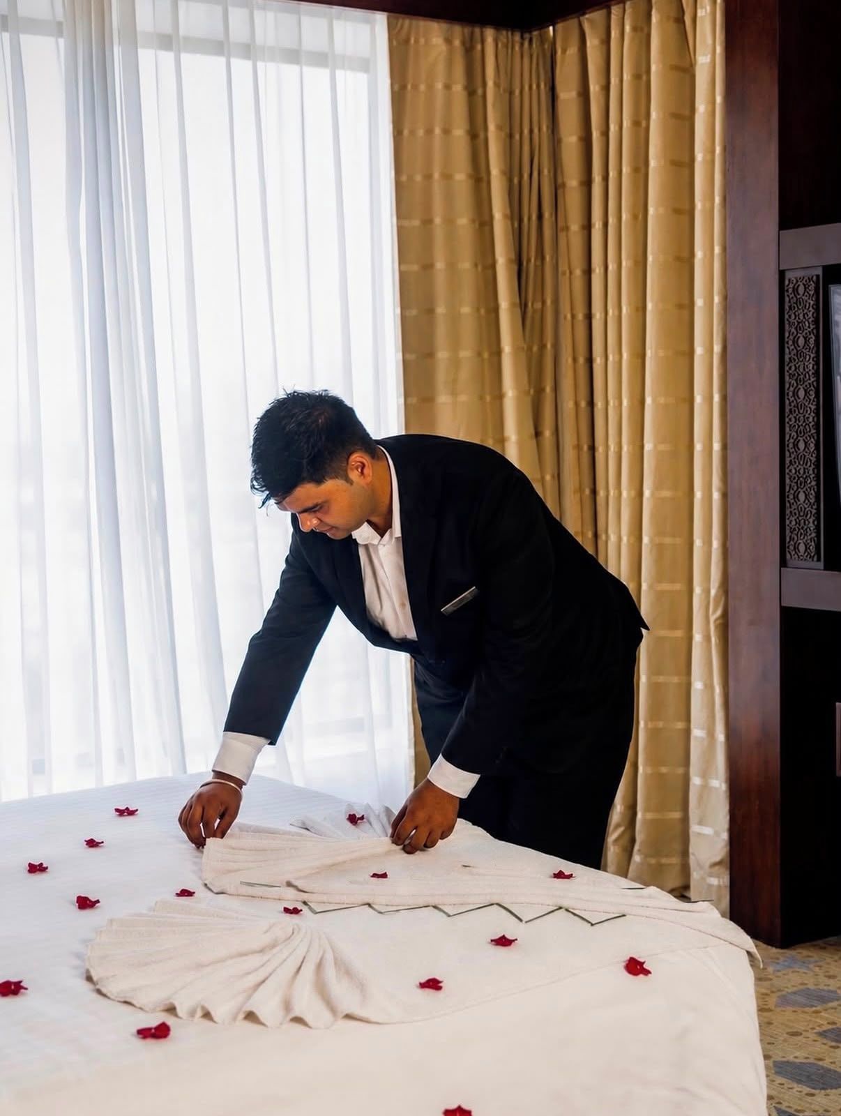 A man arranging a bed with white linens and red rose petals in a hotel room with a large window and curtains.