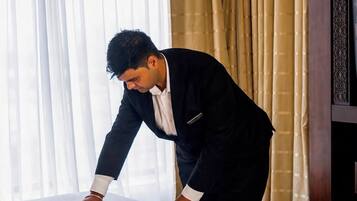 A man arranging a bed with white linens and red rose petals in a hotel room with a large window and curtains.