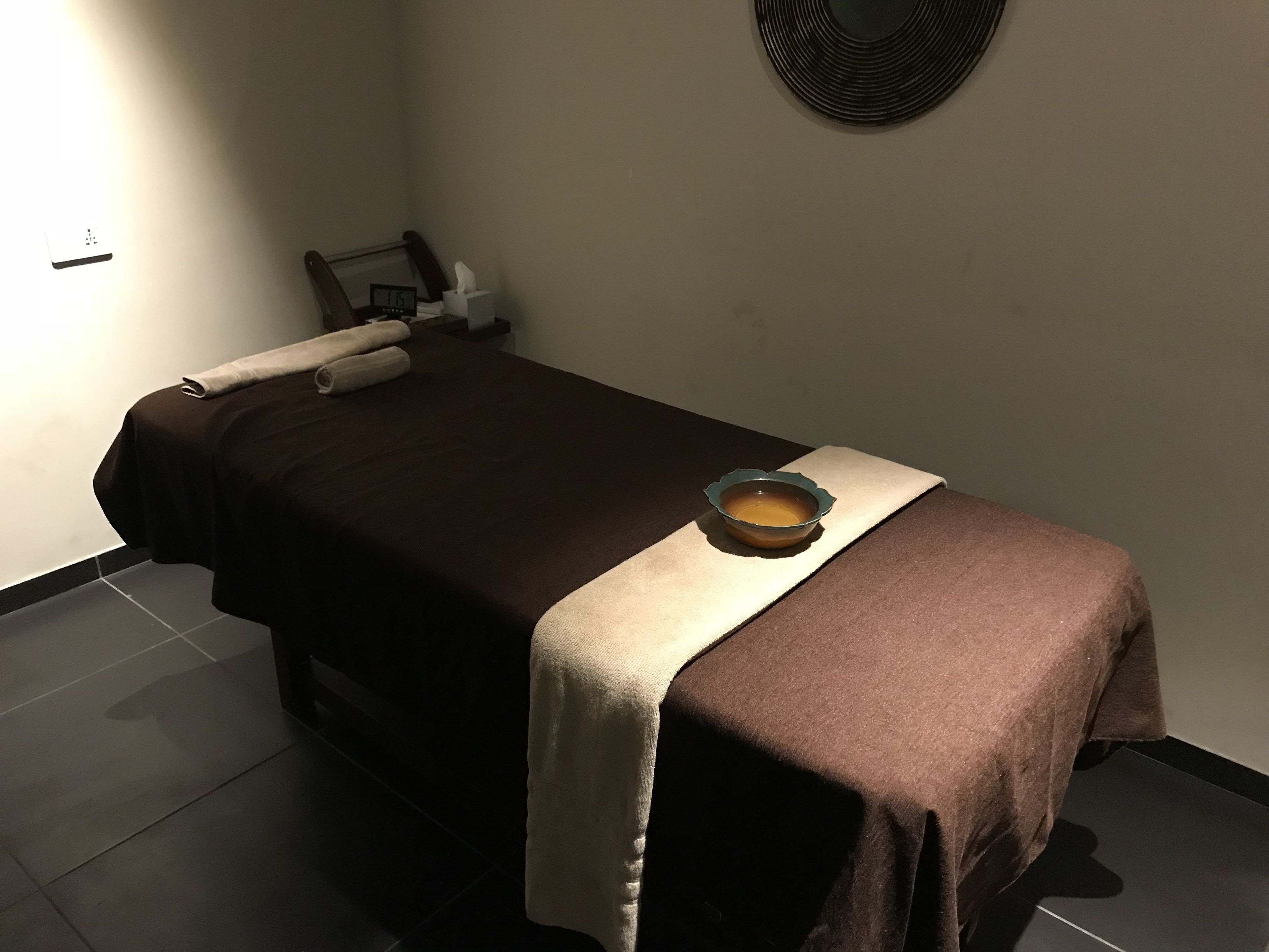 A single bed with a brown bedspread and a beige blanket, a small bowl of water, a tissue box, and a clock on the nightstand.